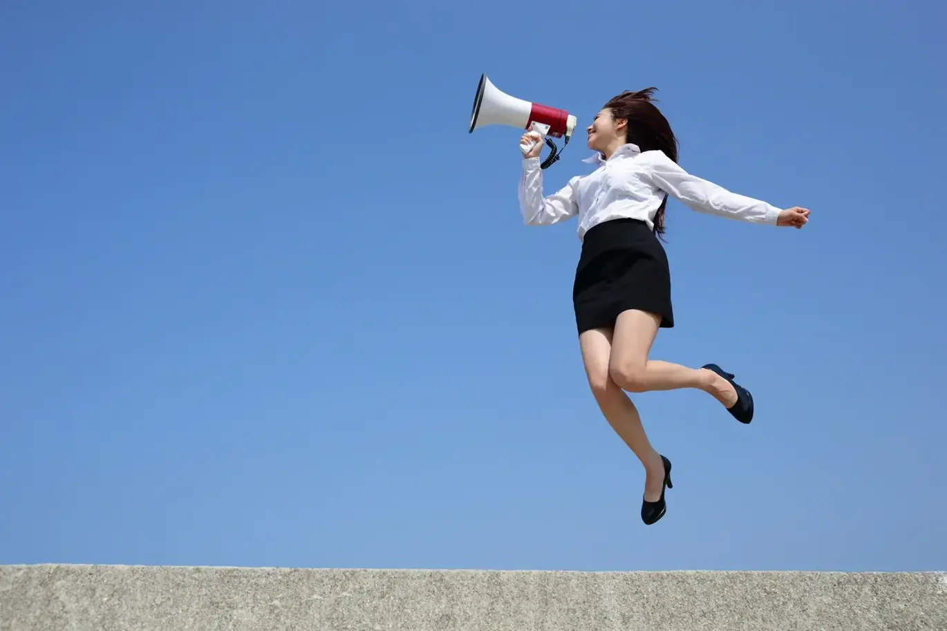 Woman jumping into air, wearing business attire and speaking into megaphone, with blue sky behind